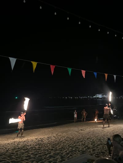 At night, at Hat Khai Muk, Thailand, fire dancers perform along a beach. The sandy beach runs along the right of the image with a few people looking on. A string of triangular flags runs from top left to bottom. A second string of lights runs parallel to the flags. The sky is very dark.