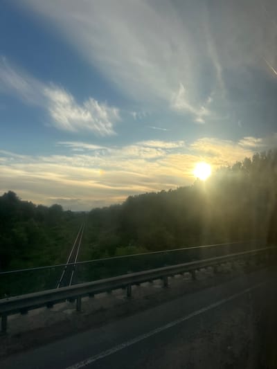 This image shows a scenic view of railroad tracks disappearing into a forest under a partly cloudy sky at sunset. The sun is low on the horizon, casting a warm glow and creating lens flare. The railroad tracks are flanked by lush green trees on either side, and a guardrail is visible in the foreground, suggesting the vantage point is from a bridge or overpass. In the distance, a faint rainbow can be seen in the sky. The location is specified as Pidbirtsi, Ukraine. There are no people or animals visible in the image.
