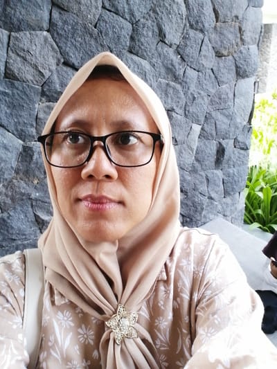 A medium shot shows a woman wearing glasses and a beige hijab. She is looking slightly off-camera to her right. She is also wearing a beige top with a floral pattern and a star-shaped brooch. The background consists of a stone wall and some greenery. The image appears to have been taken outdoors, possibly at a restaurant or cafe in Banyuwangi, Indonesia, as suggested by the stone wall commonly found in such establishments in the region.
