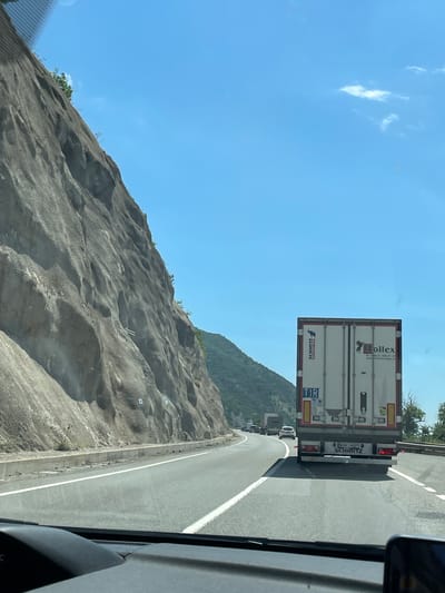 The image depicts a daytime scene from the interior of a vehicle, looking forward through the windshield onto a multi-lane highway. In the immediate foreground, a dark dashboard is visible, with reflections appearing on the windshield above.
Directly ahead, a white semi-trailer truck occupies the lane. The rear of its trailer features "SCHMITZ CARGOBULL" text vertically on the left door and a "Hollex" logo with a bull, along with a Polish phone number (+48 31 326 51 91) and website (www.hollextransport.pl), on the right door. A "TIR" plate is affixed to the left door. The truck's license plate, partially visible, displays "WSN 74492". Red taillights are illuminated.
To the left of the highway, a massive, steep rock face, grey and heavily textured, rises dramatically alongside the road. The rock exhibits vertical striations and varying contours, with small bushes and trees clinging to its surface. Visible on the rock face are multiple white markers or caps, along with what appear to be two parallel metal rods, indicating geological stabilization or monitoring. A concrete barrier separates the road from the base of the rock.
To the right, beyond the road and other vehicles in the distance, a distant, green-clad hillside is visible under a clear, bright blue sky with a few wispy white clouds. Traffic ahead appears moderate, with several vehicles visible further down the road, including another white truck and a white passenger car.
The distinctive geological features, particularly the close proximity of the road to a towering rock face, are characteristic of the DN6/E70 highway as it traverses the Iron Gates gorge near Drobeta-Turnu Severin, Romania, where the road runs alongside the Danube River through mountainous terrain.