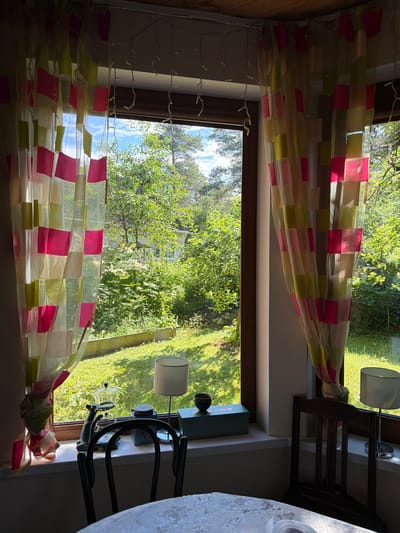 A bright, sunlit interior shot looking out a window shows a glimpse of a rural outdoor environment, possibly the city of Orekhovo, Russia, with a rustic wooden ceiling and walls. The window is adorned with sheer curtains featuring a colorful checkered pattern of pink and pale green squares. Outside, lush green trees and foliage are visible, with a hint of a white house in the background. The windowsill is lined with several objects: a small lamp, a decorative green object resembling a cat with a glass top, a blue box, and another small lamp on the right side. A dark wooden chair is positioned in front of the window, and a white tablecloth covers a table in the foreground. The overall impression is of a cozy, domestic space with a strong connection to nature.