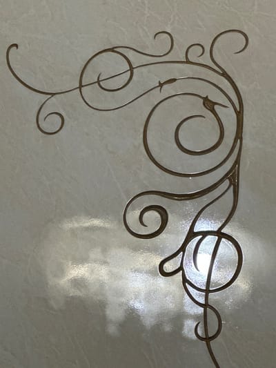 This is a close-up, overhead shot of a decorative metal scrollwork design, likely a gate or fence, in a light, textured, stone-like material. The scrollwork is dark brown and features swirling, organic shapes with sharp points and rounded curls. It appears to be an ornamental element. The background has a glossy finish, reflecting bright light, suggesting a sunny day or artificial lighting in a public space or home. The scrollwork occupies the upper left and central portions of the image, extending towards the right and downwards. The detail of the scrollwork is sharp, while the background has a softer focus due to the reflection. There is no text visible in the image. The setting appears to be an outdoor or indoor decorative element, possibly in Nookat, Kyrgyzstan, given the provided location context. The overall impression is one of ornate craftsmanship and a potentially well-lit environment.