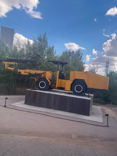 The image, taken in Satbayev, Kazakhstan, depicts a yellow drilling machine, likely a piece of industrial equipment, displayed on a concrete pedestal. The machine is oriented horizontally, with its drill arm extending outwards. The pedestal itself is decorated with a dark surface featuring a red geometric pattern. The background reveals a bright, clear sky with scattered white clouds, suggesting fair weather and daytime. Lush green trees and foliage surround the display, with a tall building visible in the distance. Power lines and a large electrical pylon are also present, indicating an outdoor, possibly urban or industrial, setting. The overall scene appears to be an exhibit or monument, showcasing the drilling equipment against a natural and constructed backdrop. No people are visible in the image, and no text is readily apparent. The lighting suggests it was taken in the late morning or early afternoon.