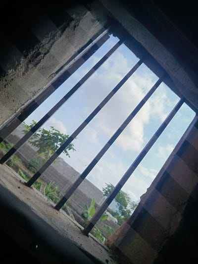 This image shows a view through a barred window looking out onto a cloudy sky and some vegetation. The window is covered with a mesh screen and has dark bars running diagonally across it. The bars are positioned at an angle, suggesting the window might be a casement or louvered window. The exterior appears to be a rural or semi-rural area, with trees and what looks like a simple building structure in the background. The concrete frame of the window is rough and unfinished. The lighting suggests it is daytime, with the sun partially obscured by clouds. There are no people or specific identifiable landmarks that would pinpoint a location in Masaka, Nigeria, or any other city.