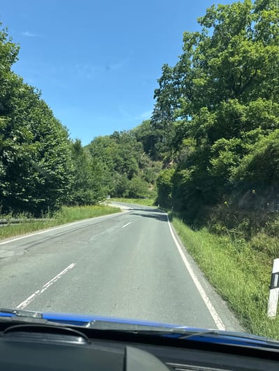 The image depicts a road in Braunfels, Germany, flanked by lush green trees and vegetation under a clear blue sky. The perspective is from inside a vehicle, suggested by the blue frame and reflective surfaces on the right side of the image. The road itself is paved and has white lane markings, indicating it's a two-way street. The time of day appears to be daytime, with bright sunlight casting shadows. The weather is sunny and clear.
The scene is characterized by natural elements. On the left side of the road, dense trees with vibrant green foliage dominate the landscape, creating a natural tunnel effect. The trees appear to be deciduous, possibly oaks, given their leafy appearance. On the right side, a grassy embankment slopes upwards, with more trees and bushes further up. A road marker, a white pole with black markings, is visible on the right side of the road, near the top of the embankment. The road curves gently to the left, disappearing into the distance.
There is no visible text in the image that provides specific information about the location or any event. The overall mood is serene and peaceful, suggesting a pleasant drive through a natural setting. The image captures a moment of travel through a picturesque German landscape, highlighting the beauty of the natural environment.