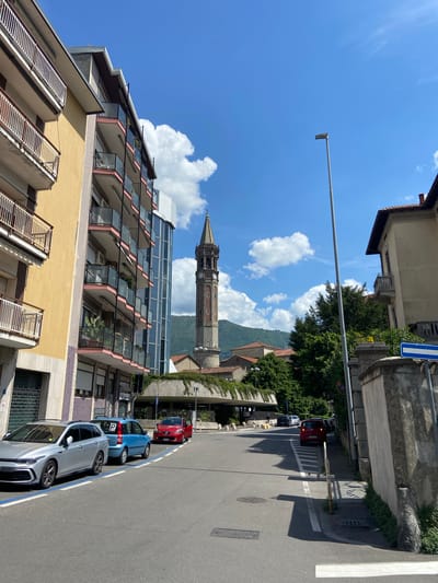 The image depicts a street scene in Lecco, Italy, under a bright blue sky with scattered clouds. A tall, ornate bell tower dominates the background, partly obscured by a modern building with glass balconies and a yellow facade. Several cars are parked along the right side of the street, including a gray, blue, and red vehicle. A street sign is visible. The road is marked with a crosswalk. In the background, a tree-covered hillside rises behind the buildings.