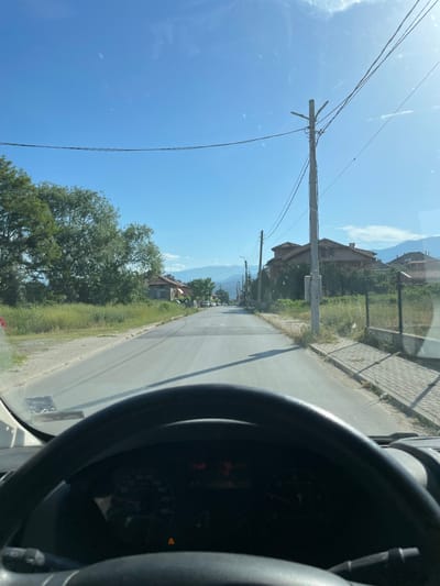 The view is from inside a vehicle, likely a car or van, looking out onto a road in Krupnik, Bulgaria. A steering wheel is prominently visible in the foreground, partially obscuring the dashboard. The road stretches ahead, bordered by greenery, houses, and a fence on the right. Power lines and poles run alongside the road on the left, reaching towards a clear, blue sky. Distant mountains are faintly visible in the background. No people are discernible in the scene.