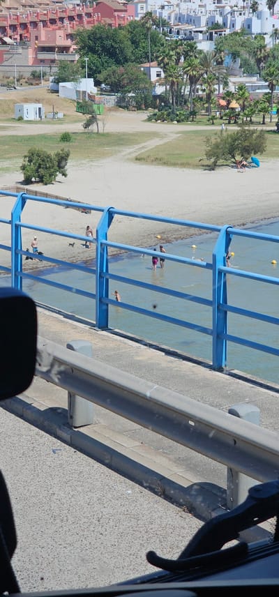 The image depicts a sunny day at a beachside location, likely Algeciras, Spain. In the foreground, a gray metal railing runs diagonally across the frame, casting a shadow. Behind it, a blue metal barrier with vertical bars separates a walkway from a body of water. Several people are visible in the shallow water, some wading and others standing.

Beyond the water, a sandy beach stretches out, lined with green grass and palm trees. Further inland, a cluster of white and pink buildings, possibly apartments or hotels, forms a dense urban backdrop. Orange and blue umbrellas are scattered on the sand. A few individuals are visible on the beach, some seated under umbrellas.

The scene appears to be a recreational area, with the blue barrier possibly part of a pier or a promenade. The bright sunlight and clear water suggest warm, pleasant weather. The overall impression is one of a relaxed summer day at the coast. There is no visible text in the image.