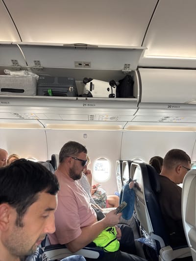 The image depicts the interior of a commercial passenger aircraft cabin, specifically focusing on the overhead bins and several seated passengers. The location is specified as ตำบล ไม้ขาว, Thailand.
In the upper left, an overhead compartment, numbered "21 CBA," is open, revealing multiple items of carry-on luggage. From left to right inside this bin: a light grey fabric soft-sided case, a dark grey hardshell suitcase with vertical ridging and a green tag that partially reads "CAE," and a white object with black accents, possibly a specialized case or another piece of luggage. A black bag is partially visible further right within the adjacent bin section. A safety decal inside the open bin displays symbols for a charging cable and a prohibited crossed-out charging cable with a battery icon. The adjacent overhead bin section to the right, labeled "20 CBA," is closed. Below the closed bin, a "Wi-Fi" symbol is affixed to the cabin wall.
In the mid-ground, a male passenger with a beard, short grey hair, and dark sunglasses is seated, wearing a pink short-sleeved t-shirt. He holds a dark-colored smartphone in his left hand and a bright lime-green fabric item, possibly a small bag or garment, in his right hand, resting on his lap. He has a ring on his right ring finger. His arm rests on the shared armrest.
To the right of the man in the pink shirt, another male passenger with short dark hair and glasses is visible from behind and in profile, wearing a dark brown long-sleeved shirt. His head is turned slightly away from the camera. A blue plush object, resembling a small travel pillow or stuffed animal, hangs from the headrest of the seat in front of him. A clear plastic bag containing an unknown item is partially visible on the floor or seat base in front of this passenger.
In the immediate foreground on the left, the left profile of a male passenger with dark hair and stubble is visible. He is wearing a patterned shirt and looking downwards. Partially visible airplane windows in the background show an indistinct external view, possibly a tarmac or sky. Overhead passenger service units with lights and vents are visible below the closed overhead bins. The aircraft seats are predominantly dark blue with light grey or white frames.