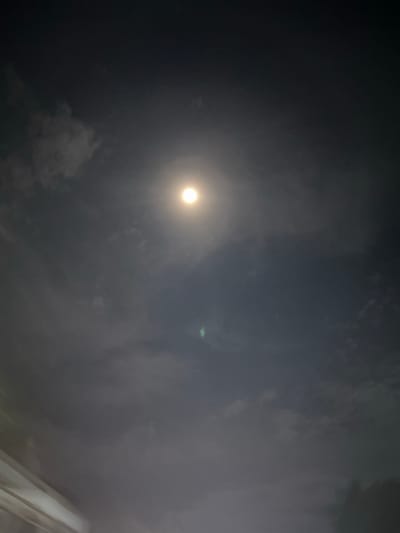 The image depicts a night sky illuminated by a bright, full moon partially obscured by clouds. The clouds are wispy and diffuse, creating a soft glow around the moon. The overall atmosphere is dark and moody, with hints of luminescence in the celestial bodies. There are no discernible human figures or man-made structures in the frame, suggesting an outdoor setting. The bottom portion of the image shows a sliver of what appears to be an artificial structure, possibly a building or a bridge, with diagonal lines indicating its form. This detail, however, is largely out of focus and does not detract from the prominence of the night sky and moon. The scene suggests a quiet, perhaps even serene, moment under the night sky, with the natural beauty of the moon and clouds taking center stage. Without further context or information about the location in Ceurih, Indonesia, it is difficult to provide specific details about the environment beyond what is visible in the image itself. The photograph appears to be taken from a ground-level perspective, looking upwards towards the heavens.