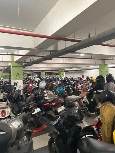 A dimly lit, multi-level indoor parking garage is filled with a large number of parked motorcycles. The environment is utilitarian, with concrete pillars and beams, and exposed red and gray conduits or pipes running along the ceiling. Green and white signage is visible on some of the pillars, indicating parking zones such as "A C-4", "A C-5", and "B-6 B-5". A sign with the word "KELUAR" (meaning "Exit" in Indonesian) is also visible. The motorcycles are of various colors and models, mostly scooters, and are parked closely together. Some motorcycles have helmets on their handlebars or seats. The lighting in the garage appears to be artificial and somewhat sparse, creating shadows. No individuals are clearly depicted engaging in any significant action, though several figures are visible in the background and foreground, many appearing to be wearing helmets and some in motorcycle gear. The overall impression is that of a busy motorcycle parking area, likely in a commercial or residential building in the city of Baji Mangngai, Indonesia.