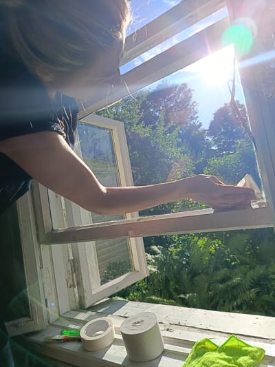 A person's arm and shoulder, wearing a dark shirt and a necklace, are visible from the side as they lean over an open window and clean the frame with a light-colored object. Outside the window, lush green trees and foliage are visible under a bright blue sky with the sun shining, creating lens flares. On the windowsill inside, there is a small paintbrush with red and green handles, a roll of white tape, and another roll of what appears to be paper towels or more tape. A bright green microfiber cloth is also on the windowsill. The window frame is old and painted white, with some peeling paint and visible hinges. The scene appears to be outdoors, possibly in a rural setting or a backyard in Niasvizh, Belarus, given the greenery and the style of the window.