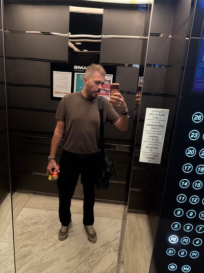 A man takes a selfie inside a modern elevator with highly reflective, dark paneling. He is seen from the chest down, wearing a brown t-shirt, black pants, and light-colored sneakers. In his right hand, he holds a red and yellow soda can and possibly a banana or yellow object, while his left hand holds a smartphone, visible as a bronze-colored iPhone, capturing the reflection. A black bag hangs over his left shoulder.

The elevator features a control panel with numerous circular buttons for different floors, labeled from 4 to 26, as well as parking levels P1, P2, P3, ground floor (G), and basement (B1). The button for "P2" is illuminated, suggesting it is either the current floor or a selected destination.

A white informational sign with both Arabic and English text is affixed to the elevator wall. It lists amenities and their corresponding levels:
- HEALTH CLUB: LEVEL 4
- SWIMMING POOL: LEVEL 4
- PARKING: P1, P2, P3
- RECEPTION: GF (Ground Floor)
- PARKING: B1

Other visible text includes a reflection of "SMART" or similar on a glass panel, and "S20" and "355" on a small card or label near the man's hand. The elevator floor is made of light-colored, marbled stone. The scene takes place indoors, within a building in Business Bay, United Arab Emirates.