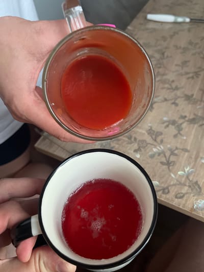 The image shows two hands holding cups of red liquid. The upper cup, a clear glass mug, contains a thick red liquid, possibly tomato juice. The lower cup, a black and white mug, contains a thinner, brighter red liquid with some foam on the surface. Both cups are held over a patterned surface that appears to be a table with a floral design. The lighting suggests an indoor setting. The image does not provide any information about the city of Navahrudak, Belarus.