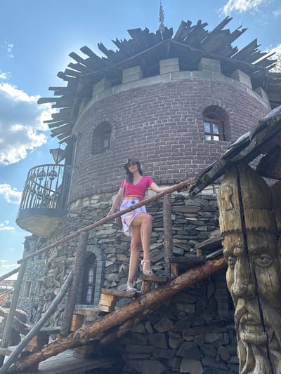 In the city of Satka, Russia, a woman is posing for a photograph on a rustic wooden staircase leading up to a stone tower. The tower, with its rough-hewn stone and brick construction, has a medieval aesthetic. The woman is wearing a pink crop top and a white and tie-dye skirt, with sandals and a hat. She is holding onto a wooden railing as she stands on the stairs. The staircase itself is constructed from rough logs and planks, adding to the rustic charm of the scene. A large, intricately carved wooden sculpture of a face stands to the right of the staircase, adding another unique element to the composition. The background features a bright blue sky with scattered white clouds, and the sun is shining, casting shadows.