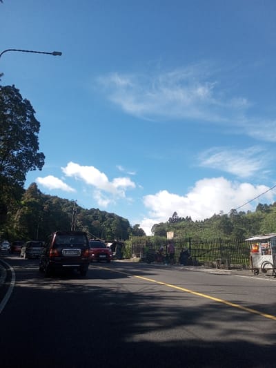 A sunny, outdoor scene depicts a multi-lane asphalt road traversing a green, hilly landscape, described as being in Ka Thaung (Upper), Myanmar. The sky is bright blue with scattered white cumulus and wispy cirrus clouds.
In the immediate foreground, a dark-colored SUV, bearing an Indonesian license plate B 3222 RLL, is positioned in the rightmost lane, facing away from the viewer. Its rear bumper and tail lights are visible. Ahead of it, a red sedan is partially visible, followed by other vehicles receding into the distance along a road that curves to the left. A prominent double solid yellow line marks the center of the road. Shadows of unseen objects or foliage are cast across the road surface.
On the far left, the dense foliage of a large tree is visible, with its upper branches extending into the frame. A black, curved street light pole with a luminaire extends from the upper left.
The right side of the image features a dark metal fence running parallel to the road, behind which a steep, heavily wooded hillside rises. Utility poles and wires are discernible against the trees. Several individuals are present near the fence line on the right: two people stand near a parked red scooter, and another individual is seated or stooping closer to a white, wheeled vendor stall with a canopy. The stall displays various items. A yellow rectangular sign is affixed to the fence above these individuals. The overall environment suggests a roadside area with commercial activity amidst natural terrain.