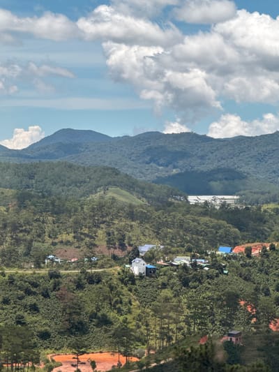 The image depicts a hilly, verdant landscape with scattered buildings and a bright blue sky dotted with white clouds. In the foreground, a patch of reddish-brown earth is visible, possibly an agricultural field or cleared land. Tall green trees, some appearing to be pine trees, are densely planted on the slopes. Several houses are nestled amongst the trees; notable are a white house with a blue roof, a cluster of buildings with blue and green roofs, and a few other structures partially obscured by foliage. Further in the background, rolling hills covered in dense vegetation recede towards the horizon. A faint suggestion of a body of water or a large, flat, illuminated area is visible in the distance between the middle and far background hills. The overall impression is one of a rural or semi-rural setting in a mountainous region. The scene appears to be located in the city of Tu Póh, Vietnam, given the architectural styles of the houses and the characteristic vegetation of the region. There are no people visible in the image.