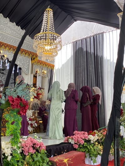 The image captures an indoor event in Matangbaroh, Indonesia, featuring an elaborately decorated space and individuals in formal attire. The ceiling is draped with black fabric, transitioning to white fabric and vertical black and white curtains on the right side. A large, ornate gold chandelier, adorned with multiple tiers of crystal-like elements and illuminated, hangs centrally from the ceiling.

To the left, a decorative stage or backdrop is prominent, featuring intricate white and gold carvings or moldings, flanked by a black fabric column. Smaller, multi-tiered chandeliers are visible in the background on the left. The foreground and lower left are heavily decorated with artificial floral arrangements, including large clusters of red and pink flowers, pink roses, white blossoms, and trailing green foliage. Fairy lights are interwoven within these arrangements on the lower right, alongside a red box-like object.

Seven individuals, all appearing to be women, are present. On the far left, a woman in a purple outfit and matching hijab is partially obscured by foliage. On the stage, a young girl or woman in a purple outfit and hijab is seated on an ornate grey upholstered chair, looking towards the right. In the midground, a woman in a light mint green long dress and matching hijab stands with her back partially to the viewer, facing right. To her right, a group of four women stands together, appearing to converse. From left to right within this group, one woman wears a light brown hijab and purple attire, another wears a dark maroon hijab and dress while holding a dark handbag, a third wears a light beige hijab and a reddish-brown outfit and is smiling, and the fourth wears a light beige hijab and a reddish-brown dress, mostly facing away from the camera. All visible individuals wear headscarves (hijabs) and modest long dresses or kebayas.