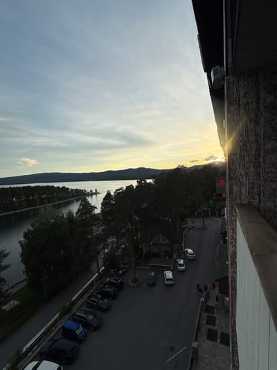 The image shows a view from an elevated position, possibly a balcony, in Dom Otdykha Zolotoy Plyazh, Russia. A body of water stretches into the distance, reflecting the sky. Trees line the shoreline and a road runs parallel to the trees, with several parked cars visible. On the other side of the road are various buildings. In the distance, the sun is setting, creating a bright glow. The sky is filled with clouds. The balcony or building structure is visible at the top and bottom of the frame.