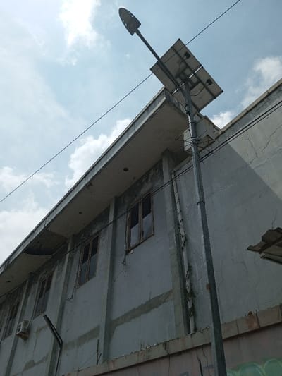 The image displays a low-angle view of a street scene in Viengthong, Laos, under a partly cloudy daytime sky. A tall, gray metal streetlight pole stands vertically on the right, supporting a rectangular solar panel array near its top. Above the solar panels, a dark, elongated LED street light fixture extends outward. Multiple black utility wires traverse the frame, connected to the pole and extending across the upper portion of the image.

To the left and behind the streetlight pole is a multi-story concrete building with a visibly weathered light-gray facade featuring extensive cracking, discoloration, and dark stains. A prominent concrete overhang or balcony ledge is visible across the upper section of the building. Below this ledge, a row of multiple rectangular windows with dark frames is visible on an upper floor. On the far left, an external air conditioning unit is mounted on the building wall beneath a window. Faint green and black graffiti markings are present on the lower section of the building's wall, partially obscured. No individuals are present in the scene.