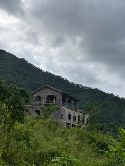 A stone building with multiple arched windows and balconies is situated on a hillside in Lùng Tám Commune, Vietnam. The building's exterior is constructed from rough-hewn stones, giving it a rustic appearance. On the upper balcony, there are several figures visible, suggesting human presence. The building is surrounded by dense green foliage, including trees and tall grasses. In the background, a steep, tree-covered mountain rises, shrouded by dramatic, cloudy skies. The overall scene depicts a building integrated into a lush, natural environment under overcast conditions.