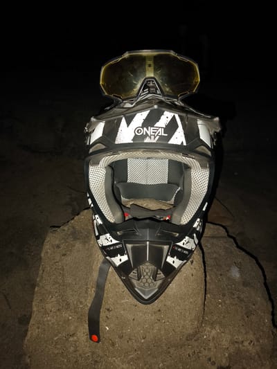 A black and white Oneal motocross helmet with yellow tinted goggles rests on a concrete structure outdoors. The helmet is predominantly black with white geometric patterns and the brand name "ONEAL" prominently displayed. The goggles are positioned on top of the helmet. The helmet strap hangs down on the left. The background is dark, suggesting a nighttime setting. The concrete structure the helmet is resting on appears weathered. No people are visible. The location cannot be confirmed as Huanchaco, Peru, based on the visual information provided.