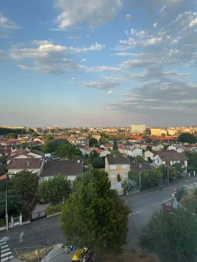 The image captures an aerial view of a residential area in Colomiers, France, during what appears to be late afternoon or early evening, indicated by the warm lighting and soft shadows. The foreground is dominated by the rooftops of numerous houses with terracotta tiles, interspersed with lush green trees and vegetation. A street runs through the scene, with a red car visible on it. Further in the distance, the cityscape unfolds with various buildings, including a prominent white multi-story building and industrial structures, suggesting an urban or semi-urban environment. The sky above is a clear blue, dotted with scattered white and grey clouds. The scene depicts a quiet suburban neighborhood with elements of urban sprawl in the background.