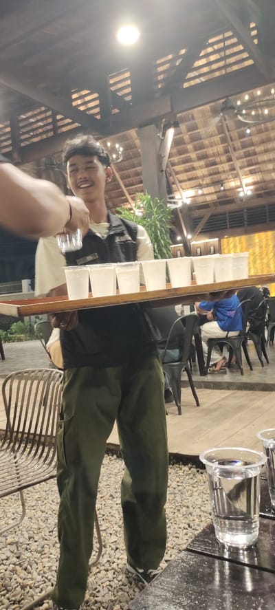 A young man, likely a waiter, is holding a wooden tray with several clear plastic cups filled with a clear liquid. He is wearing a dark vest over a light-colored shirt and olive green cargo pants. His facial expression is a wide, happy smile, and he appears to be in motion, possibly serving the drinks.
In the foreground, a person's arm and hand are visible, reaching towards the tray, perhaps to take a cup or assist the waiter. Another tray with two more cups of liquid is on a dark surface in the lower right of the frame.
The setting appears to be an outdoor or semi-outdoor dining area with a rustic aesthetic. A wooden roof with exposed beams is visible overhead, illuminated by bright lights. There are decorative string lights hanging from the ceiling. In the background, other patrons are seated at tables, some blurred, and there are metal chairs and tables. A lush green plant is also visible. The ground is covered in small, light-colored stones.
The scene likely takes place in the evening, given the artificial lighting. The atmosphere appears casual and friendly, indicated by the waiter's joyful expression and the relaxed setting. The location context suggests Bireuenmeunasahdayah, Indonesia. There is no visible text in the image.