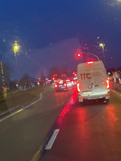 The image depicts a nighttime or dusk scene from the interior of a vehicle, looking forward through a wet windshield, characterized by numerous water droplets and streaks. The location is in the city of Evergem, Belgium.

The scene features a multi-lane road, wet from precipitation, with vehicle traffic. Directly in front, occupying the right lane, is a white commercial van with its red taillights and high-mounted stop light illuminated, indicating it is stationary or moving slowly. The van displays a "ttc" logo in red text on its rear right door, with smaller, unreadable text below it. A circular sticker is present on the lower left rear door. Reflections of red vehicle lights and traffic signals are prominent on the wet asphalt road surface.

Ahead of the white van, multiple other vehicles are visible, primarily as blurred clusters of red taillights, also appearing to be stopped. At least one blue passenger car is discernible behind the van. Overhead, multiple red traffic signal lights are illuminated, indicating a stop condition. Yellow-orange streetlights illuminate the scene from both sides of the road.

On the left side of the road, a paved area or sidewalk is visible, with indistinct shapes possibly representing pedestrians or stationary objects. A tall, yellow-and-black striped pole is situated on the far left edge of the frame. The sky is dark blue, consistent with evening or night conditions. No specific individuals or their actions are clearly identifiable.