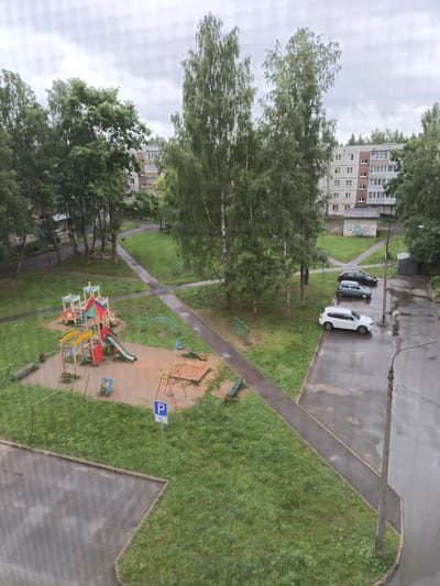 This aerial view captures a courtyard scene in Tikhvin, Russia, under an overcast sky, suggesting either early morning or a cloudy day. The ground is wet, indicating recent rainfall. In the foreground, a children's playground features a colorful climbing structure with slides, swings, and a sandbox. A blue and white parking sign with a wheelchair symbol is prominently displayed near the playground, designating accessible parking.
To the right of the playground, a paved driveway curves along the edge of the grassy area. Several cars are parked on the wet pavement, including a white SUV, a light blue hatchback, and a dark sedan. A concrete light pole stands on the right side of the frame, with wires extending from it.
In the background, apartment buildings with multiple stories and windows are visible, interspersed with tall, leafy trees, characteristic of a residential area. A narrow paved path winds through the courtyard, connecting different areas. The overall impression is a typical urban residential courtyard, likely in a post-Soviet era housing complex, with play areas for children and parking facilities for residents. No people are visible in the image.