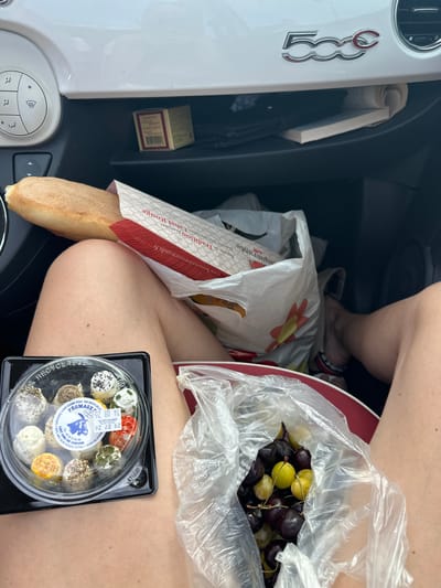 The image shows a close-up view of groceries on the lap of a person sitting in a car. The groceries include a baguette in a paper bag, a container of small cheeses, and a plastic bag filled with grapes. The car's interior is visible, with the dashboard and glove compartment in the background. The image appears to have been taken in or around Ouville-la-Rivière, France, based on the information on the cheese packaging, which indicates "Fromagerie Saint Cosme SCEA - 600 Route de Béthy," a location in Saint-Cosme-de-Vair, France, and a "LAIT CRU DE CHEVRE" (raw goat's milk) label. The specific location within Ouville-la-Rivière is not ascertainable from the image alone.