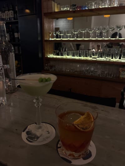 In a dimly lit bar, two cocktails are prominently displayed on a marbled countertop. The cocktail on the left is in a stemmed martini glass and appears to be a pale green hue, garnished with three green spherical objects on a skewer. To the right, a second cocktail, a darker amber color, is served in a rocks glass and is garnished with a slice of dried orange. Behind the bar, shelves are lined with various bottles of liquor and an assortment of glassware, illuminated by warm accent lighting. The overall ambiance suggests a sophisticated establishment, possibly a cocktail bar in Taito, Japan.
