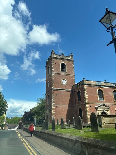 The image depicts St. John's Church in Knutsford, United Kingdom, from a low-angle street view. The red brick church tower with a clock face is visible against a bright blue sky with white clouds. A portion of the main church building is also visible, along with gravestones in the adjacent churchyard. A lamp post is in the upper left corner. A road with double yellow lines is in the foreground. A person in a red coat is walking on the pavement. Buildings and trees are visible in the background.
