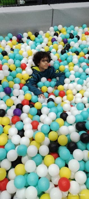 A child with curly hair lies in a ball pit. The pit is filled with white, turquoise, yellow, red, black, and purple plastic balls. The child wears a dark jacket with a yellow design on the shoulders. The ball pit appears to be indoors, with padded grey walls visible on the left side.