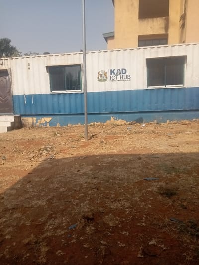 The image depicts an outdoor scene in Kaninkon, Nigeria, under clear daylight conditions. The central subject is a horizontally oriented shipping container, repurposed as a building, featuring a white upper section and a blue lower section with noticeable paint peeling. A coat of arms is prominently displayed on the white section, alongside the dark text "KAD ICT HUB" and the smaller slogan "EMPOWER. INNOVATE. EXCEL." Two dark-framed windows with security grates are visible on the container's face. To the left, a brown, paneled wooden door is set back slightly, accessed by a single concrete step. A tall, slender, light-colored metallic pole stands vertically in the foreground, positioned in front of the container's right half. The ground consists of reddish-brown earth, largely bare with scattered patches of dry grass and small debris. A distinct, dark shadow occupies the lower portion of the foreground. In the background, behind the container, a multi-story building with a light beige or yellow facade and dark windows is partially visible beneath a clear, light blue sky. No individuals or active interactions are present in the scene.