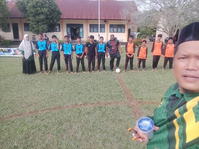 In the city of Blangriek, Indonesia, on a grassy field in front of a school building, a group of young males in athletic uniforms are posing for a photograph. A woman in a white hijab and dark clothing stands on the left, and a man in a patterned shirt is on the right, with a soccer ball at his feet. The school building has a reddish-brown roof and light-colored walls with visible windows and doorways. The field is marked with dirt lines, suggesting a sports area. In the foreground, a person wearing a green and yellow uniform holds a clear plastic cup and a pen. The overall scene depicts a casual gathering, possibly before or after a sports event.