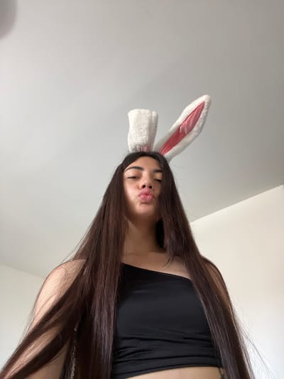 This is a low-angle, wide shot of a young woman wearing a fluffy white bunny ear headband with pink inner lining. Her dark hair cascades down past her shoulders, partially obscuring her body. She is wearing a black top. Her head is tilted back slightly, and her eyes are closed as she puckers her lips in a kissy-face pose. The background is a plain, light-colored wall, suggesting an indoor setting. The lighting is bright and even, with no discernible shadows that would indicate a specific time of day or weather conditions. The scene is simple and focused on the woman's playful expression and the bunny ears. There is no visible text in the image. The location context is Chotěšov, Czechia.