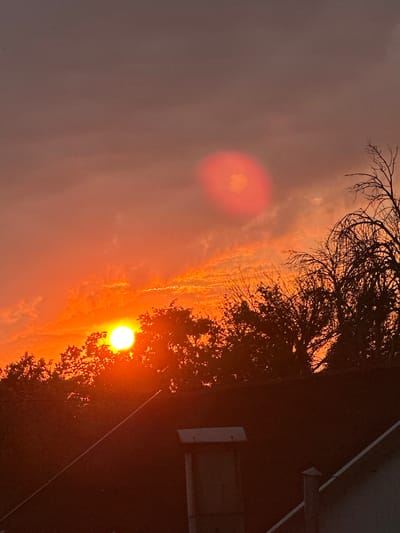 The image shows a sunset over a residential area, likely in Primorsko-Akhtarsk, Russia. The sun is a bright orb of orange, partially obscured by the branches of a tree on the right side of the frame. The sky is filled with dramatic clouds, colored in shades of orange, pink, and purple, indicating a vibrant sunset.
In the background, a dark, sloped roof of a building is visible, with a white line running diagonally across it. There are also hints of a white rectangular object on the side of the building, possibly a light fixture or a vent. The overall scene suggests a peaceful, late afternoon or early evening setting. The lighting is warm and atmospheric, evoking a sense of calm and natural beauty. The camera angle is tilted, giving a dynamic perspective to the sunset and the surrounding elements. No people or text are visible in the image.