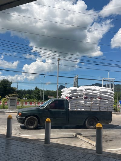 In Tambon Phraek Ha, Thailand, a dark green pickup truck is parked outdoors. The truck's bed is heavily loaded with numerous white sacks, which appear to be tied down with ropes. In the foreground, there are two cylindrical bollards with yellow caps, situated on a tiled surface. The truck is parked on a paved area with white lines, suggestive of a parking lot or roadway. In the background, there are trees, utility poles with wires, and a cloudy sky with patches of blue. Some buildings and mountains are also visible in the distance.