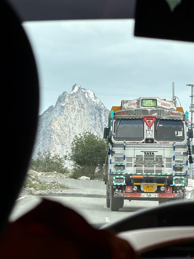 The image shows a front view of a white Tata truck adorned with decorative elements and signage. The truck is on a paved road, with a prominent snow-capped mountain range in the background. The sky is overcast. The scene appears to be in a mountainous, rural area. The truck's license plate is visible and reads "HP 72 C 9457". The truck has "TATA" written on its grille, and above the windshield, it has "GOODS CARRIER" signage. There are also various colorful decorative stickers and symbols on the truck's exterior. The perspective suggests the photo was taken from inside another vehicle.