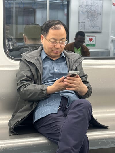 An Asian man wearing glasses, a blue button-down shirt, a gray vest, and navy pants is lying on a gray surface, possibly a seat, and looking at a cell phone he is holding. His feet, in black shoes, are positioned against a vertical support. In the background, a man with dark skin is visible, as well as a map and logo. The scene is likely set in the Tiaodeng, China subway car.