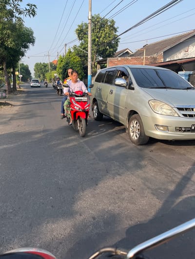 In Sugih Waras, Indonesia, a silver van is parked on the right side of the road. In the foreground, a person is riding a red motorcycle, with another person behind them. The motorcycle is moving forward, passing the van. In the background, other motorcycles and a white car are also visible on the road. The scene is set on a sunny day with a clear blue sky. The road is paved, and there are trees and houses on either side.