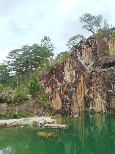 This is a landscape photograph of a quarry lake in Lán Tranh, Vietnam. The lake's surface is a vibrant, deep green, reflecting the surrounding environment. A small, wooden raft is visible floating on the water near the shore. The quarry walls are steep and rocky, displaying a striking pattern of stratified rock in shades of brown, orange, and grey. Vegetation, including various trees and shrubs, grows along the edges and on top of the quarry. In the background, a dense forest of tall, slender pine trees lines the upper rim of the quarry. The sky is overcast, with soft, diffused light. There are no people present in the scene.