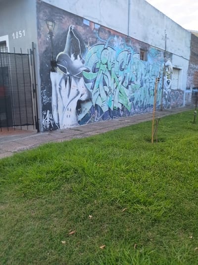 The image shows a graffiti-covered concrete wall in El Talar, Argentina (latitude: -34.462, longitude: -58.652). The wall is painted with a mix of green, blue, and white spray paint, forming abstract shapes and lettering. A large, white, cartoon-like figure is depicted on the left side of the wall, seemingly wearing a hat and tie. The graffiti is located next to a concrete sidewalk and a small patch of grass, with a metal fence and a gated entrance to the left of the wall. The wall has a visible street number, "1851". A broken light fixture is attached to the wall, and there is a small tree growing in the grass near the edge of the sidewalk.