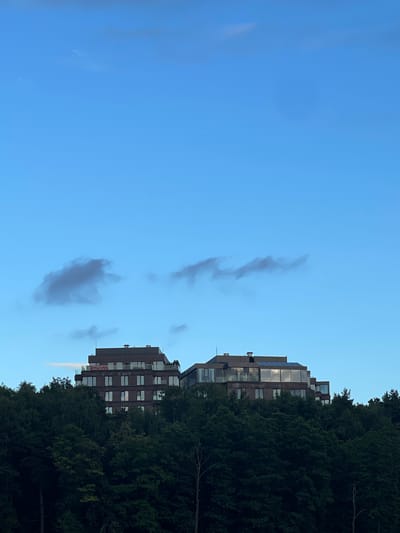 This media file captures a scene of modern architecture nestled amidst dense greenery, with a clear blue sky overhead. The image is oriented vertically, with the sky occupying the left half and the buildings and trees on the right.

The main subjects are two multi-story buildings, constructed with a contemporary design featuring large windows and balconies. They appear to be residential or office complexes, with one building exhibiting a sign that reads "ПРОДАЕТСЯ" (For Sale) in red lettering. The buildings are partially obscured by a thick canopy of dark green trees, suggesting a location where nature and urban development coexist.

The sky is a vivid blue, with a few wispy clouds scattered across it. The lighting suggests it is daytime, likely late afternoon or early evening, given the soft light and the shadows cast by the trees. The overall mood is tranquil and serene, highlighting the blend of man-made structures and the natural environment.

There are no people visible in the frame. The activity taking place is not immediately apparent, but the "For Sale" sign indicates a commercial aspect related to the property. The location context provided is Perepechino, Russia, which helps to place the scene geographically.