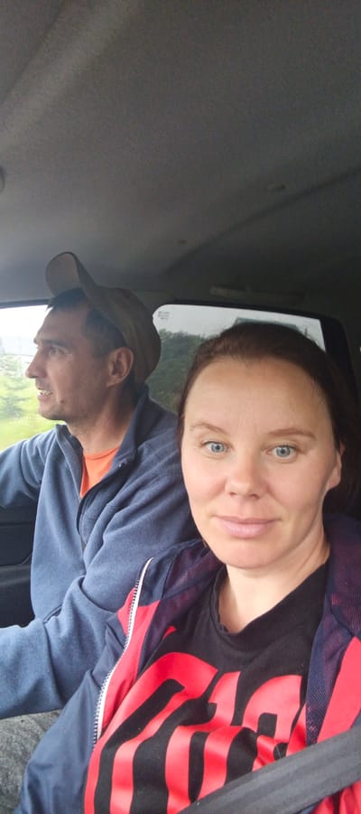 This media file is a selfie taken inside a vehicle, likely a truck, in Yakshur-Bod'ya, Russia. The primary subjects are a man and a woman, both appearing to be in their 30s or 40s.

The man is seated in the driver's seat, wearing a blue hooded jacket over an orange t-shirt, and a beige baseball cap. He is looking towards the left of the frame, with his mouth slightly open as if speaking or mid-sentence.

The woman is in the passenger seat, closer to the camera. She has light brown hair and striking blue eyes. She is wearing a black t-shirt with a prominent red graphic print and a navy and red jacket zipped up. She is looking directly at the camera with a relaxed, pleasant expression.

The setting is the interior of a vehicle, with the dashboard and ceiling visible. Through the windows, we can see greenery and trees, suggesting an outdoor, possibly rural, environment. The lighting indicates it is daytime, likely overcast or slightly hazy, as there are no harsh shadows.

The activity appears to be a casual journey, possibly a road trip or a commute. The overall mood is relaxed and friendly, with both individuals seemingly comfortable and engaged with each other and the selfie. There is no visible text in the image.