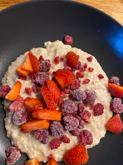 A close-up shot depicts a black plate on a wooden surface. The plate holds a serving of oatmeal, topped with chopped strawberries and raspberries. The berries appear to be partially frozen, indicated by a frosty coating. The scene does not contain people or actions, only the presentation of food. It is not possible to determine if the image was taken in Lommedalen, Norway, or any specific location, from the visual information provided.