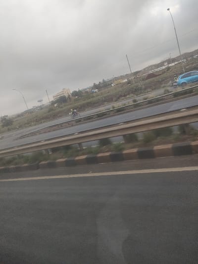 The image presents an outdoor highway scene in Gatongora ward, Kenya, captured under an overcast sky. The foreground shows a dark asphalt road surface, delineated by a solid yellow line and a curb featuring alternating black and brown painted sections. A metal guardrail runs across the mid-ground, separating the viewer's perspective from the multi-lane highway beyond.
On the highway, a single individual operates a motorcycle, dressed in a bright yellow reflective vest, proceeding leftward on one of the inner lanes. Further to the right, a light blue hatchback car is stationary on the road's shoulder, facing left. A second individual, wearing a light-colored vest, stands near the driver's side front fender of the blue vehicle. The highway incorporates multiple lanes and a central, vegetated median strip.
In the background, an elevated embankment, sparsely covered with vegetation, is visible. It features prominent white text advertising "Rockside Paints." Beyond the embankment, several structures are present: a multi-story building with a light-colored facade on the left, and a cluster of industrial-style buildings with light roofs to the right. Various utility poles and streetlights are positioned throughout the distant landscape.