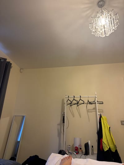 The image shows an interior wall featuring a crystal chandelier, a two-tiered shelf, and a mirror. The chandelier, with a silver base, is affixed to the left wall. On the opposite wall, a white metal shelf holds various items, including clothes, containers, and a small lamp. A mirror with a white frame is positioned near the bottom of the wall. The location is Swindon, United Kingdom.