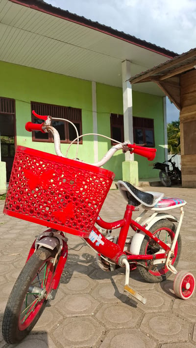 A bright red child's bicycle with training wheels is parked on a light-colored hexagonal paver patio in front of a light green building with a brown roof. The bicycle has a red basket adorned with butterfly cutouts on the handlebars. The building appears to be a school or community center, with multiple windows visible. To the right of the building, a portion of a wooden structure is visible. A black motorcycle is parked in the background. The sky is cloudy.